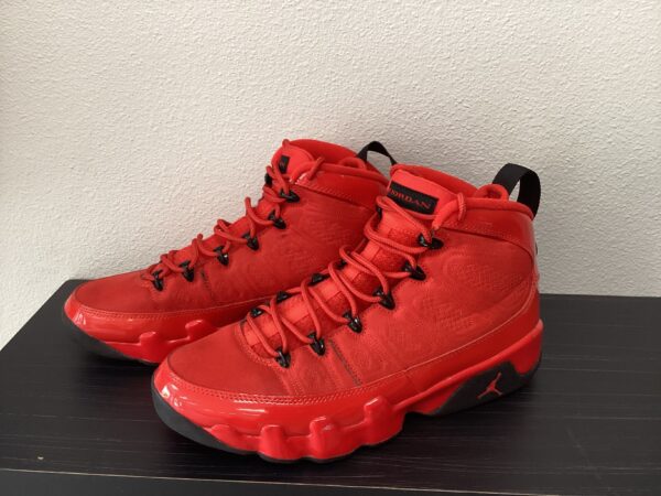 A pair of bright red pre-owned Jordan 9 basketball shoes with black accents, displayed on a black table against a white wall.