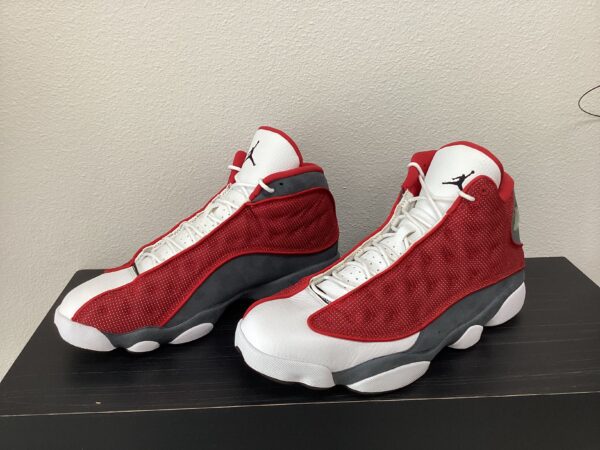 A pair of red and white Pre-Owned Jordan 13 sneakers displayed on a black surface against a white wall.