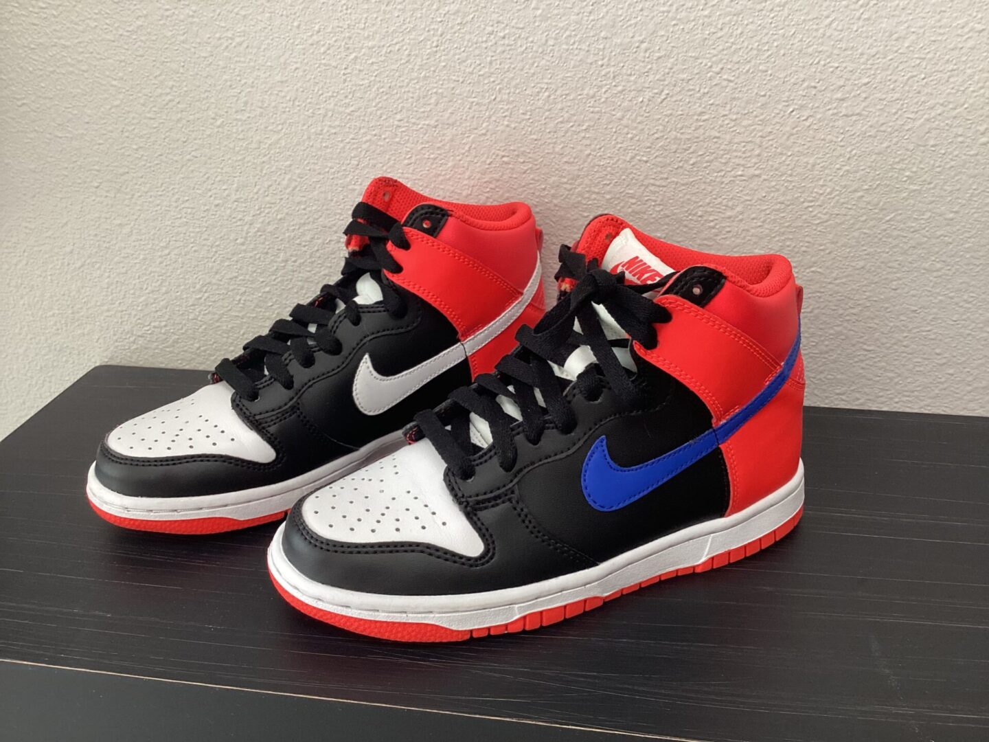 A pair of pre owned Dunk High Tops sneakers with black, red, and white colors and blue accents, displayed on a dark shelf against a gray background.