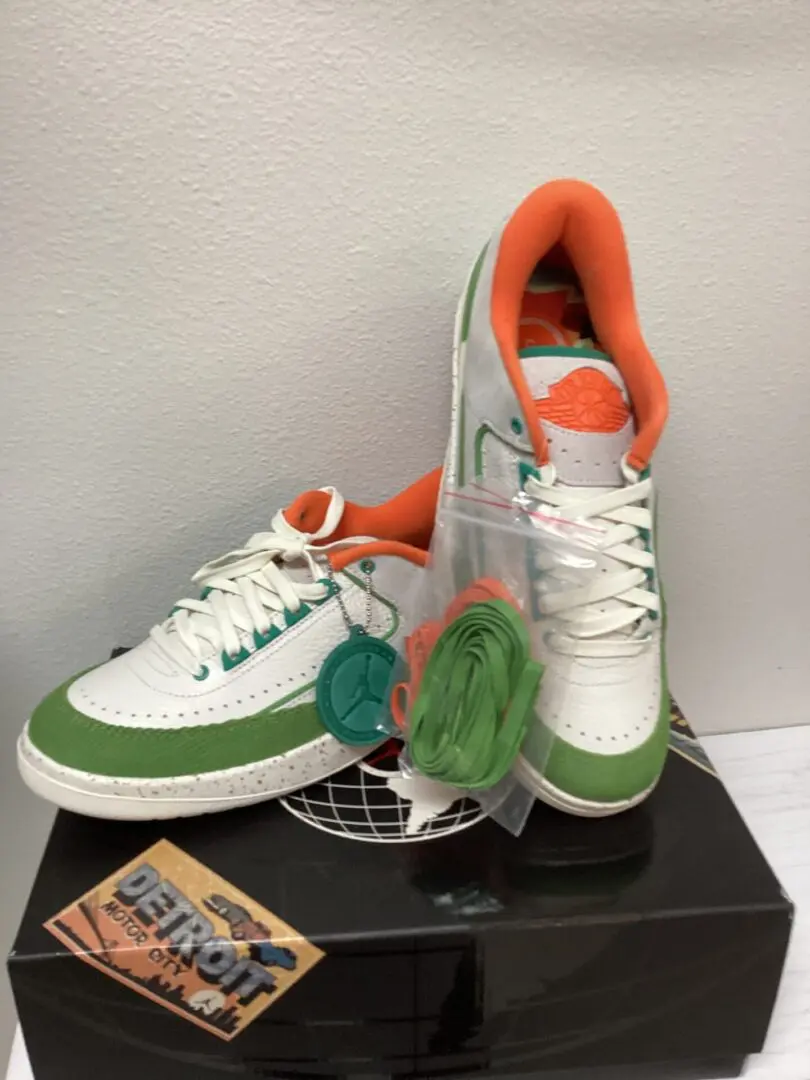 A pair of Jordan 2 Retro Low sneakers in green and white with additional laces attached, displayed on a black pedestal.