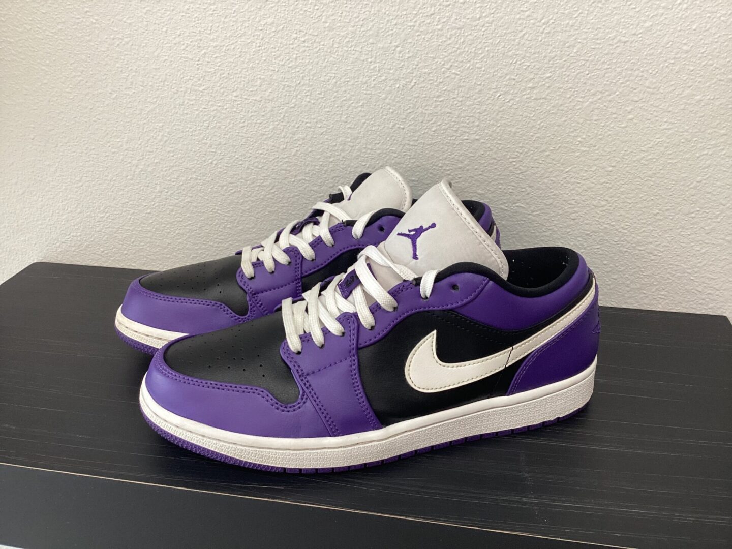 A pair of purple and black sneakers with white laces and a white Pre owned-Jordan 1 Low swoosh logo on a gray shelf.