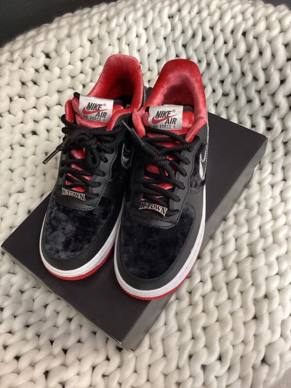 A pair of black Nike Air Force 1 Low sneakers with red interiors, placed on a white woven rug.