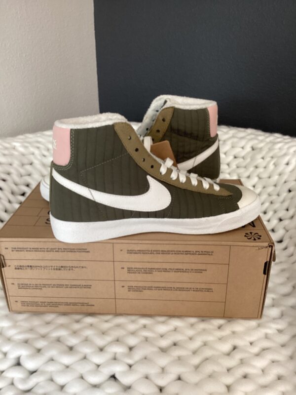 A pair of Nike Dunk Low Retro high-top sneakers in olive green and pink displayed on a shoebox, set against a white textured background.