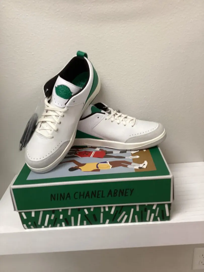 A pair of new Jordan 2 Low-Nina Abney Green/White sneakers with green accents on a shoebox featuring colorful artwork, displayed on a white shelf.