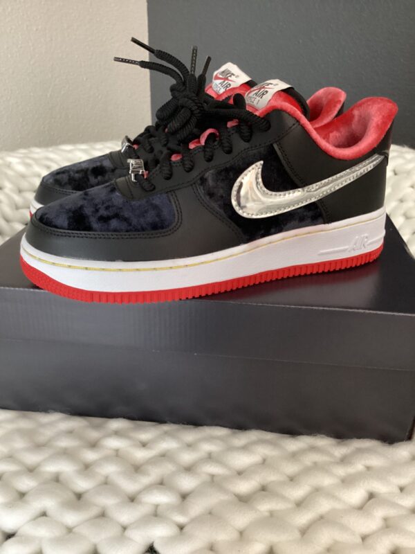 A pair of black Air Force 1 Low sneakers with red soles and a fuzzy exterior, displayed on a black box against a white textured background.