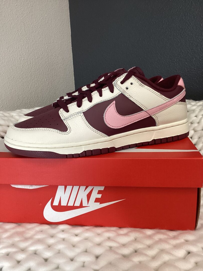 A pair of maroon and white Nike Dunk Low Retro sneakers displayed on top of a matching shoe box, set against a plain, light-colored backdrop.