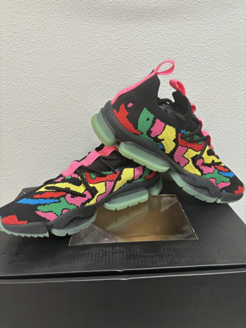 A pair of Sia Collective MACHE Multicolor sneakers with abstract patterns displayed on a light gray surface against a white wall.
