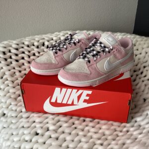A pair of pink W Nike Dunk Low sneakers placed on top of their red shoebox on a white knitted blanket.