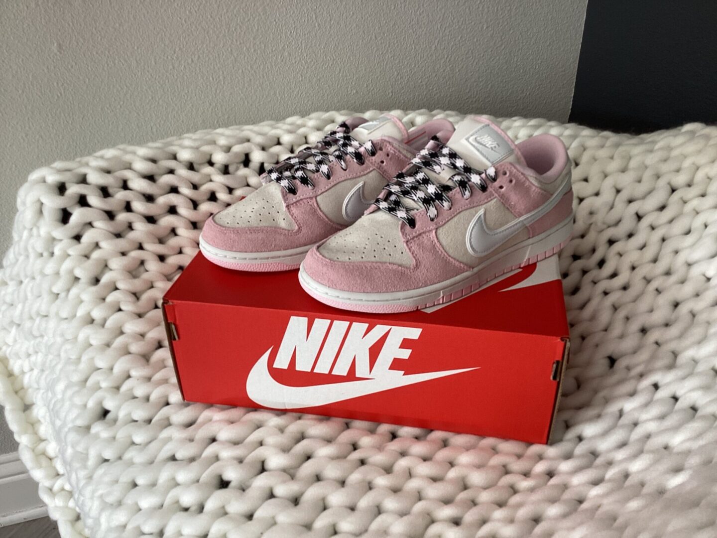 A pair of pink W Nike Dunk Low sneakers placed on top of their red shoebox on a white knitted blanket.
