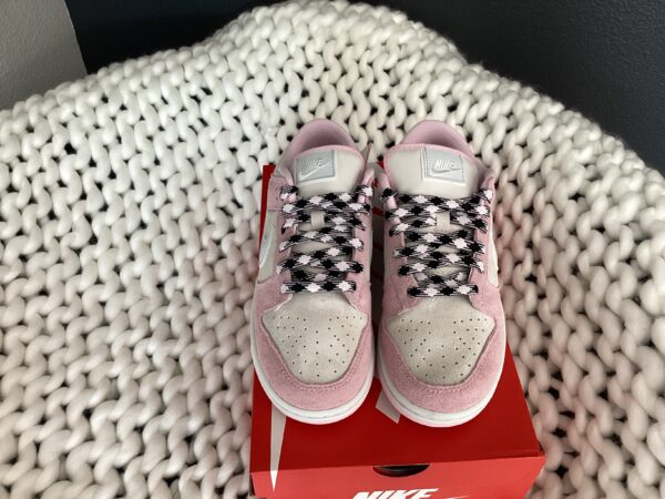 A pair of pink W Nike Dunk Low sneakers with black and white laces, placed neatly on top of their box, set against a white textured blanket.
