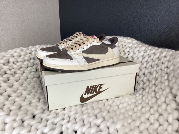 A pair of Nike Air Jordan 1 (Travis Scott) sneakers placed on top of a shoebox on a textured gray rug.