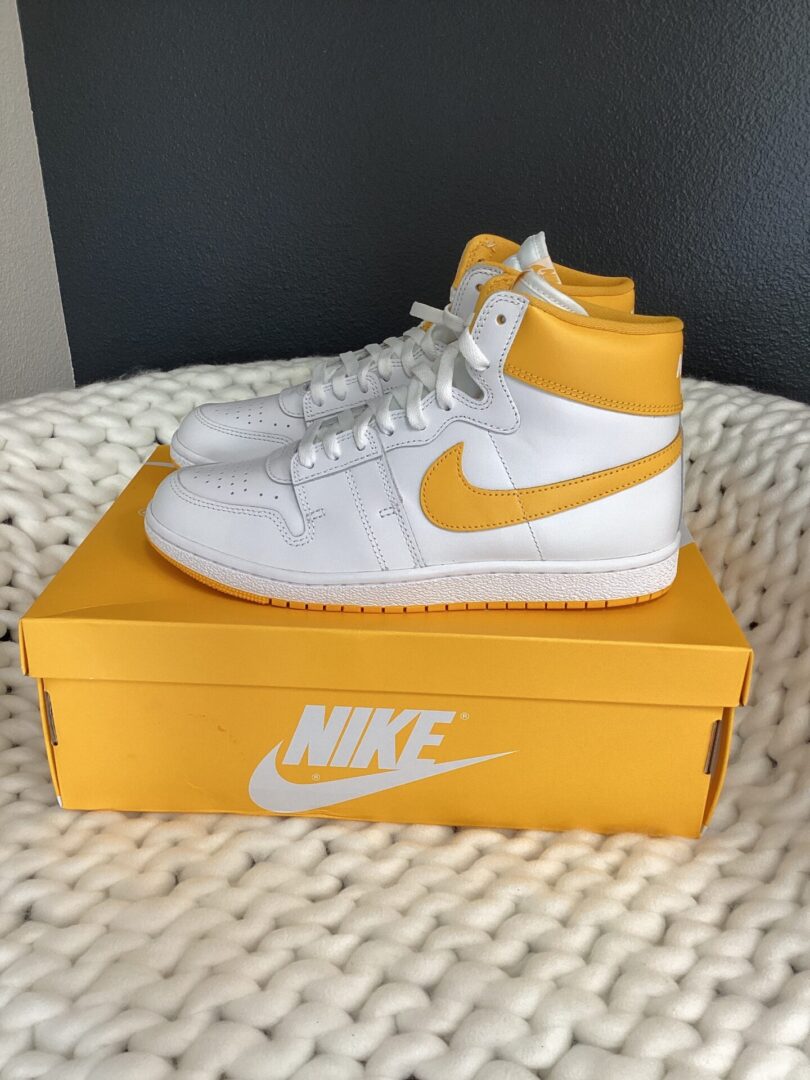 White and yellow nike air jordan 1 sneakers displayed on top of their shoe box, placed on a textured white blanket.