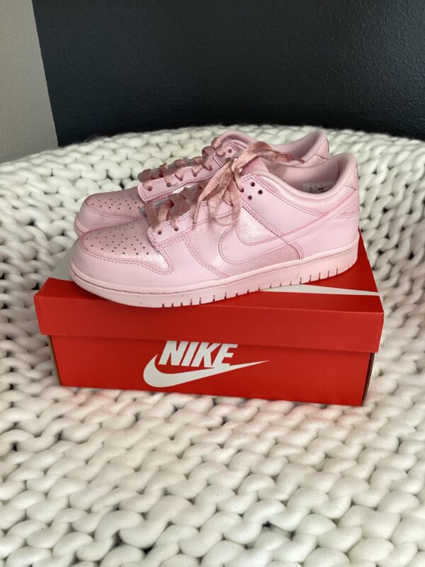 A pink Nike Dunk Low displayed on top of its red shoebox, sitting on a white textured surface.