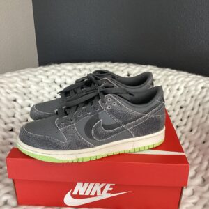 A pair of gray Nike Dunk Low sneakers with a green sole, displayed on top of a red Nike Dunk Low shoebox, on a white textured surface.
