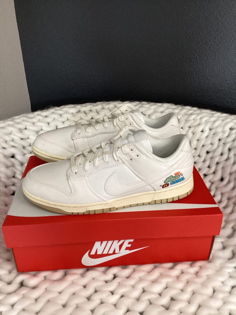 Pair of white Nike Dunk Low sneakers with a small colorful logo on the side, displayed on a Nike shoe box, against a grey wall and white textured surface.