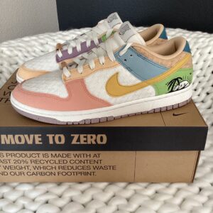 A pair of colorful Nike Dunk Low Low (Sun Club) Sail/Sanded Gold sneakers on a shoebox labeled "move to zero," placed on a white textured surface.