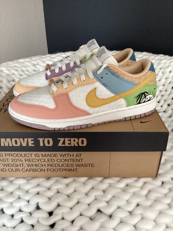A pair of colorful Nike Dunk Low Low (Sun Club) Sail/Sanded Gold sneakers on a shoebox labeled "move to zero," placed on a white textured surface.