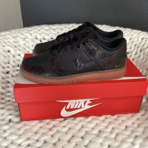 A single black Nike Dunk Low Black/Velvet Brown sneaker displayed on top of its red Nike shoebox, placed on a white textured surface.