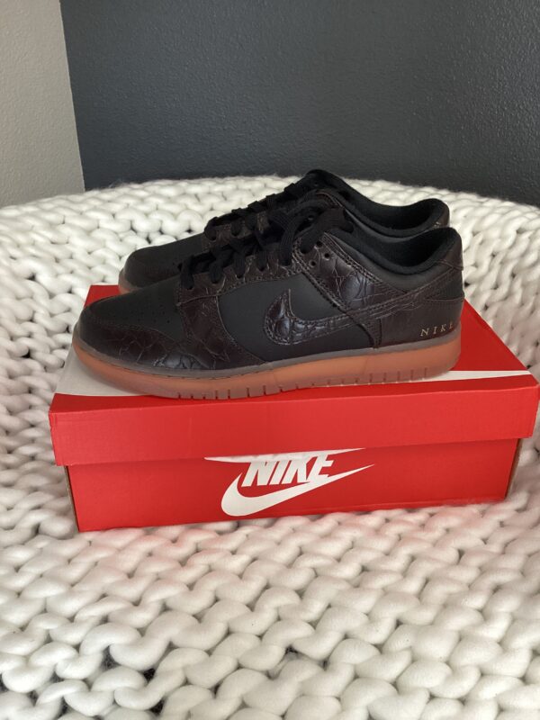 A single black Nike Dunk Low Black/Velvet Brown sneaker displayed on top of its red Nike shoebox, placed on a white textured surface.