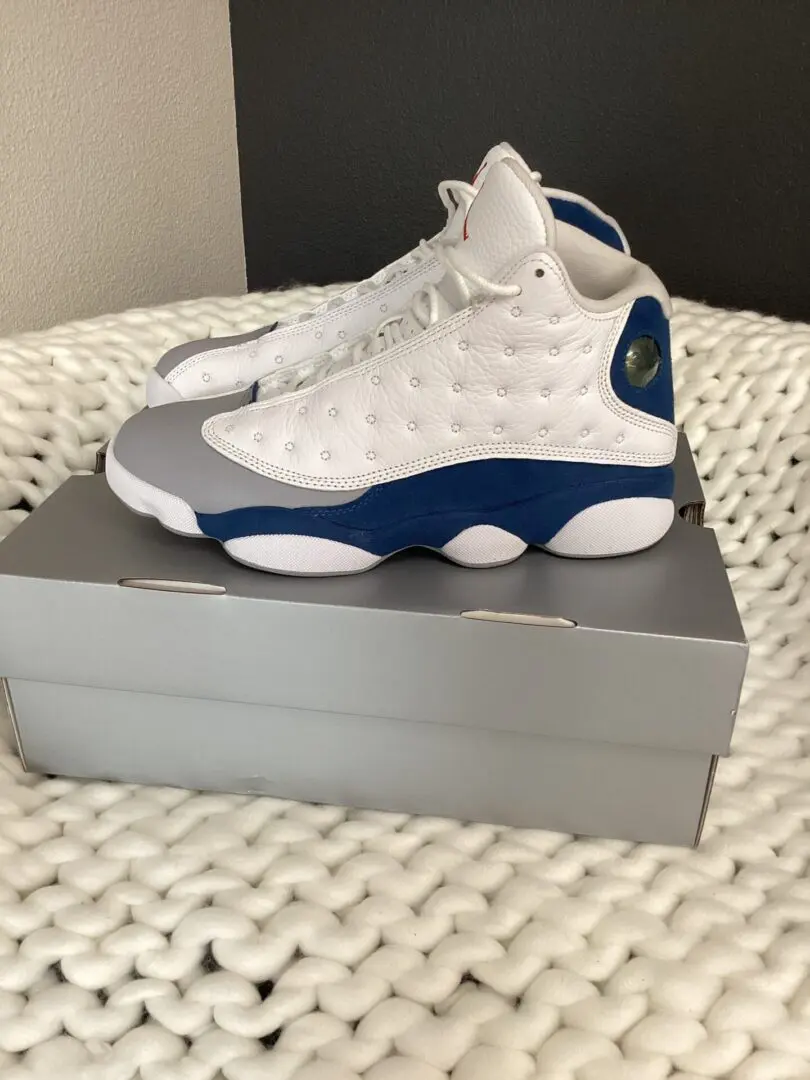 A pair of white and blue Jordan 13 French Blue sneakers on top of a gray shoebox, placed on a white textured rug.