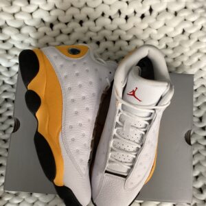A pair of white and yellow Jordan 13 Del Sol sneakers arranged neatly on top of their shoebox, placed on a textured white knit surface.