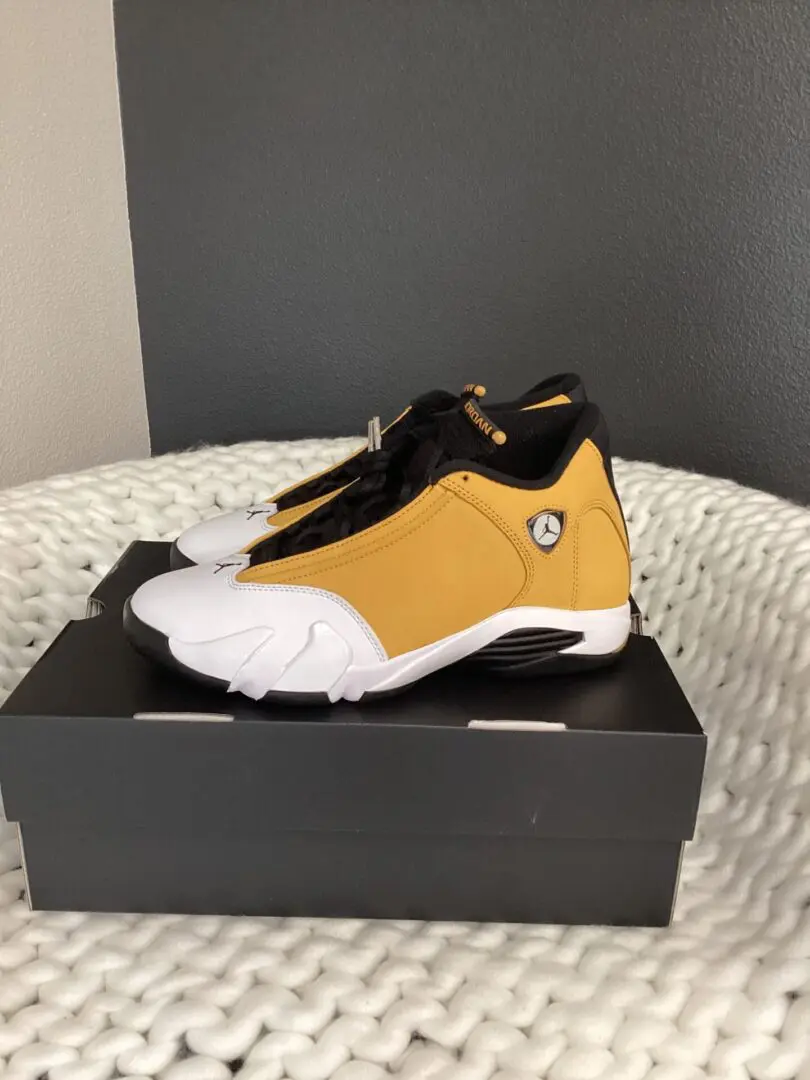 A pair of white and yellow Jordan 14 sneakers displayed on a black box set against a gray textured backdrop.