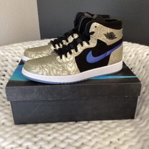 A pair of black and gold Jordan 1 Comfort Laser Gold sneakers with a blue nike swoosh, displayed on a shoe box, placed on a white textured surface.