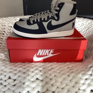 A pair of white and navy blue Nike Terminator High sneakers displayed on a red Nike shoebox, placed on a white textured blanket.