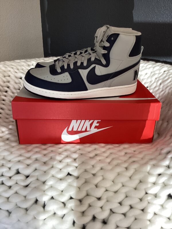 A pair of white and navy blue Nike Terminator High sneakers displayed on a red Nike shoebox, placed on a white textured blanket.