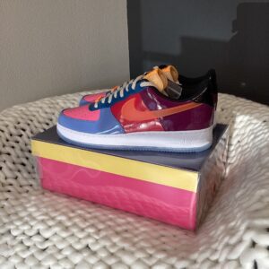 A colorful Air Force 1 Low Multi Color sneaker with blue, pink, and red panels, displayed atop its branded shoe box on a white textured surface.