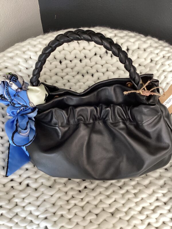 A black leather Burberry Bucket Bag with a braided handle and a blue and white patterned scarf tied to one side, resting on a white textured cushion.