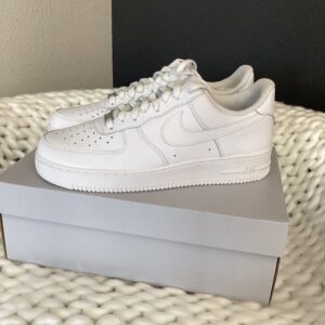 Sentence with replaced product name: Nike Air Force 1 White sneakers displayed on a shoebox, resting on a textured white blanket against a grey wall.