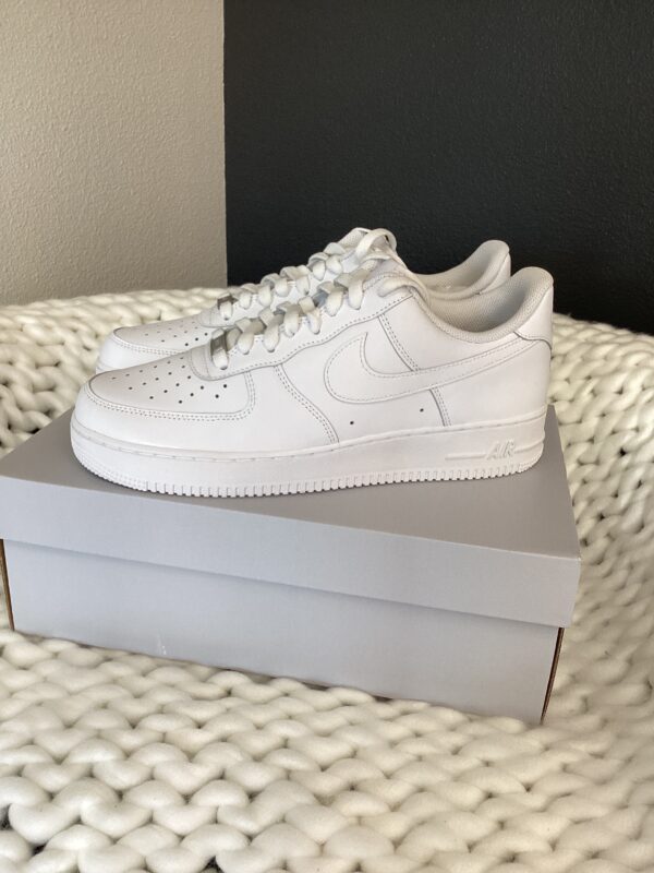 Sentence with replaced product name: Nike Air Force 1 White sneakers displayed on a shoebox, resting on a textured white blanket against a grey wall.