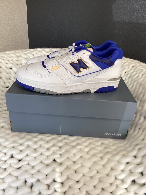 A pair of New Balance 550 White/Blue sneakers with blue and yellow accents, displayed on their box, which is placed on a textured white rug.