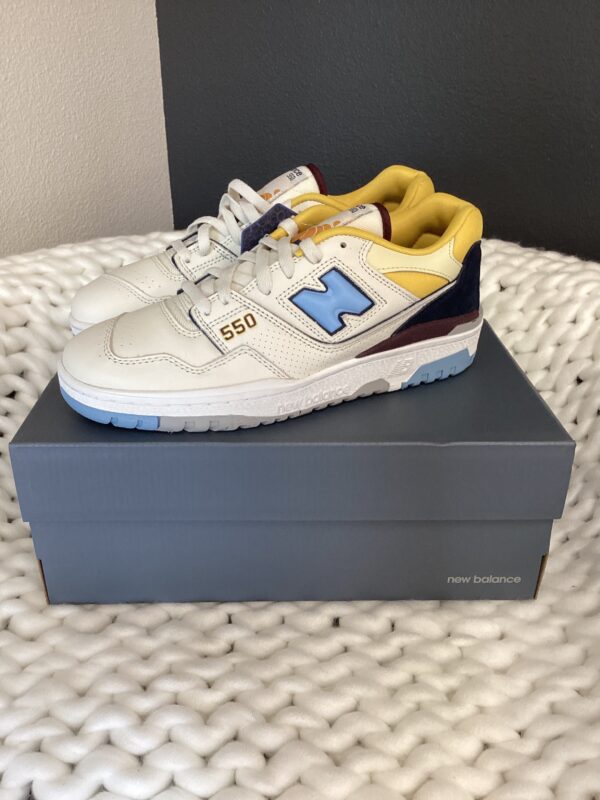 Sentence with replaced product: A pair of New balance 550 White/Yellow/Blue sneakers, displayed on their box set on a textured white surface.