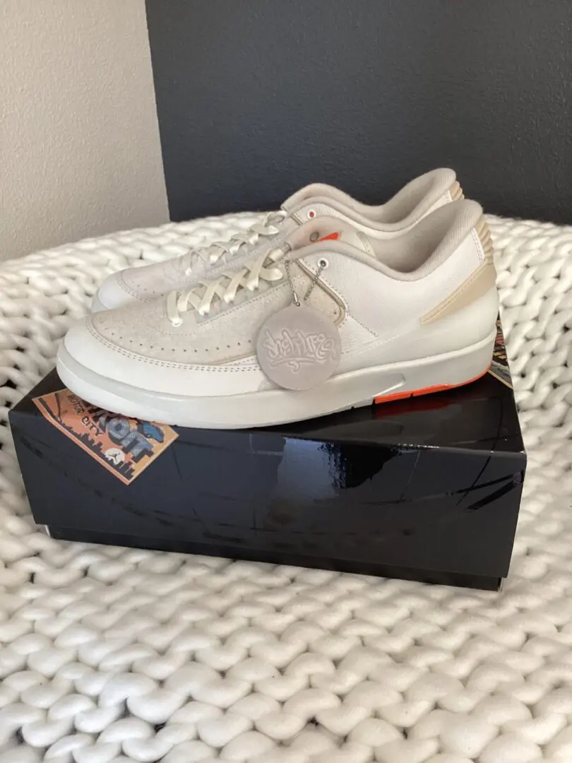 A pair of Air Jordan 2 Retro sneakers with grey accents and orange details, displayed on a shoebox, placed on a white textured surface.