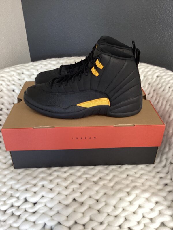 A pair of black Air Jordan 12 Retro sneakers with gold accents displayed on top of their original box, placed on a woven white surface.