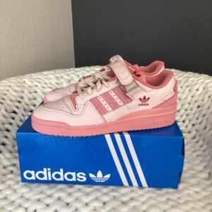A pink Adidas Forum Pink sneaker displayed on top of its shoebox, which is placed on a textured white surface.