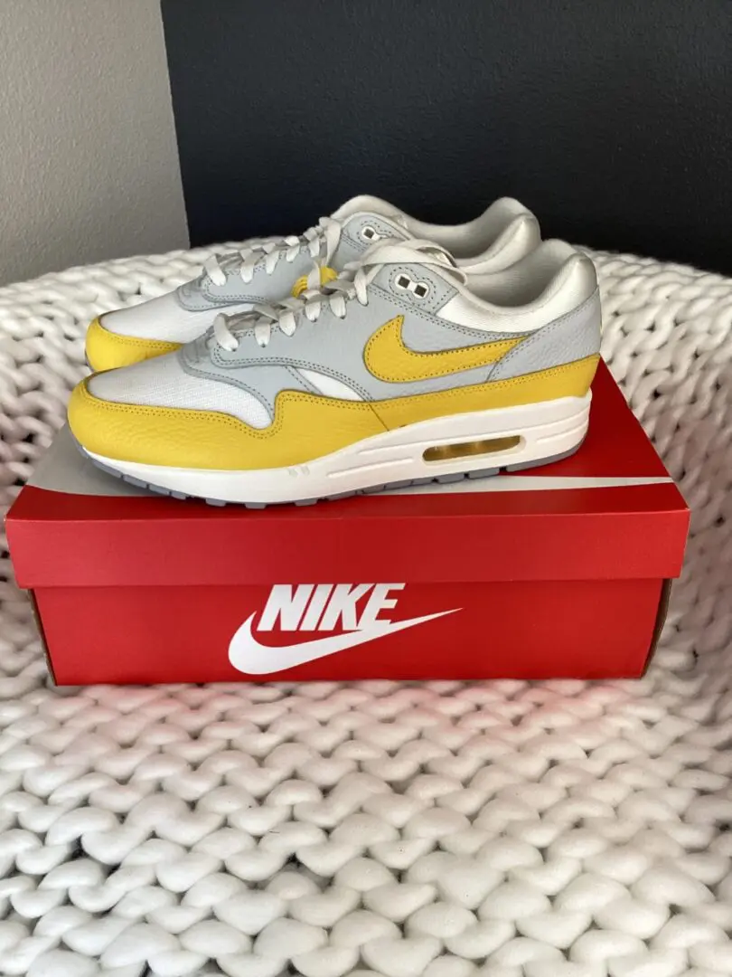 A pair of gray and yellow Sourced Air Max 1 sneakers displayed on top of a red Sourced Air Max 1 shoebox, placed on a white textured surface.