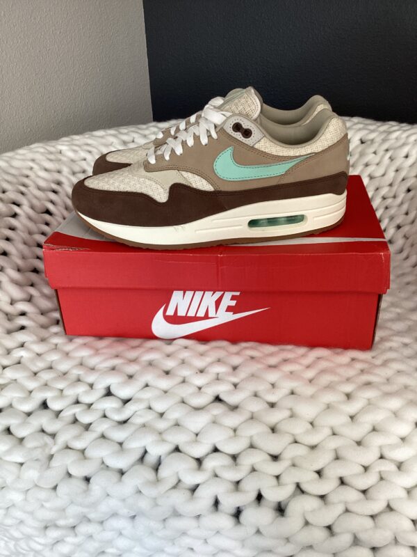 A pair of Air Max 1 Brown Mint Foam sneakers, with a teal swoosh, displayed on a red nike box on a white textured surface.