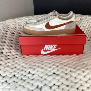 A pair of white Killshot 2 Leather sneakers with orange swoosh logos placed on top of their red shoebox, set on a white textured blanket.