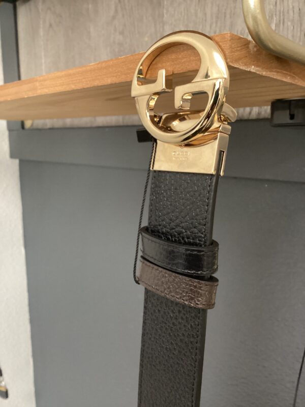A Gucci Black/Brown Belt with a gold-tone buckle hanging on a wooden rack against a gray wall.