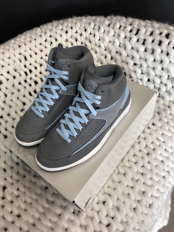 A pair of gray Air Jordan 2 Retro sneakers with light blue laces, displayed on top of a shoebox, against a white knitted background.
