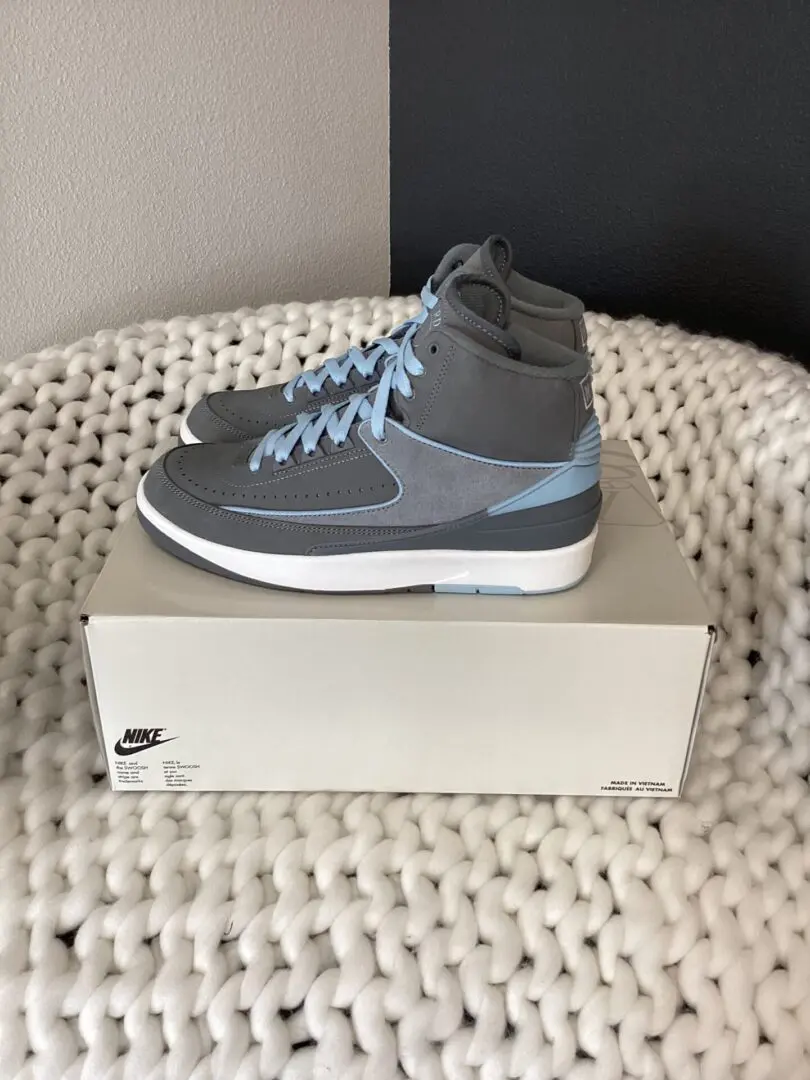 A pair of grey Air Jordan 2 Retro sneakers with light blue laces, displayed on top of their shoebox, placed on a white textured surface.