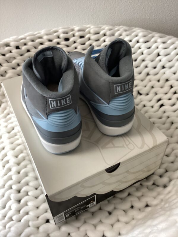 A pair of grey Air Jordan 2 Retro sneakers placed on their box, which is resting on a white knitted blanket. the shoes have blue accents and the nike logo visible.