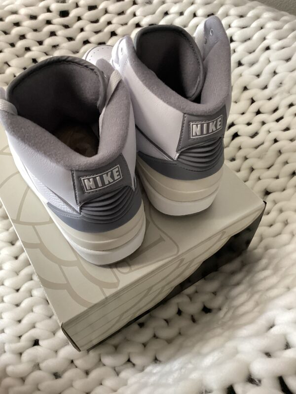 A pair of gray and white Air Jordan 2 Retro sneakers placed neatly on top of a shoe box, set against a white textured background.