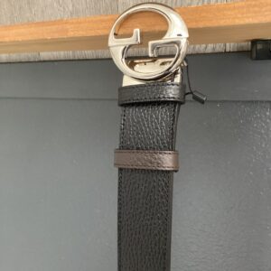 A Gucci black leather belt with a silver buckle hanging from a wooden rod against a grey textured background.