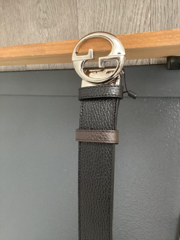 A Gucci black leather belt with a silver buckle hanging from a wooden rod against a grey textured background.