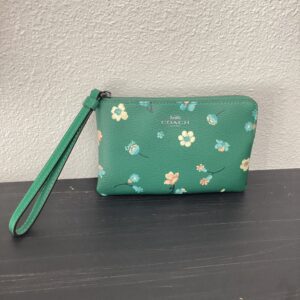 A small coach wristlet with a floral pattern on a teal background, placed on a gray surface against a white wall.