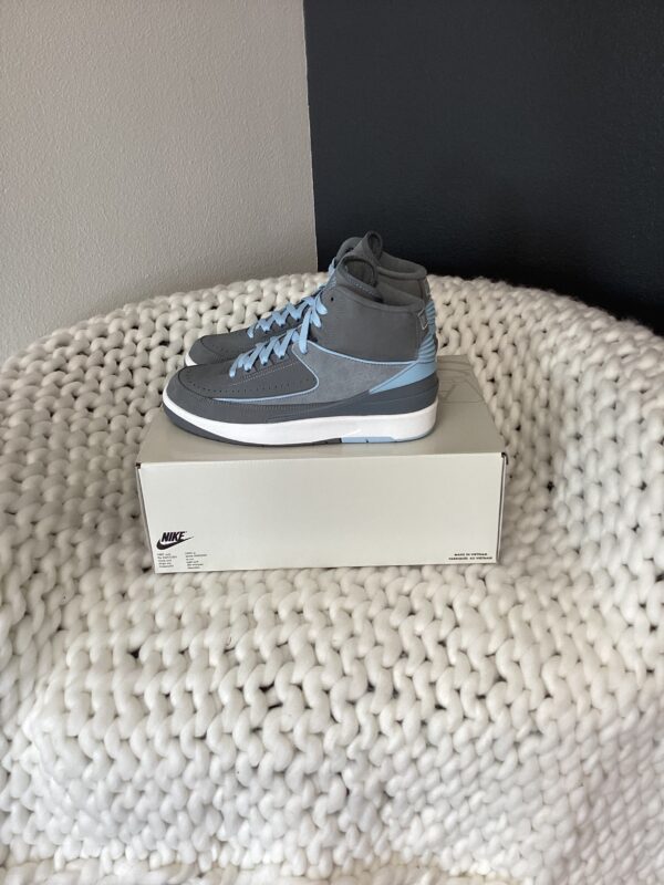 A gray Air Jordan 2 Retro sneaker displayed on its shoebox, placed on a textured white knitted surface.